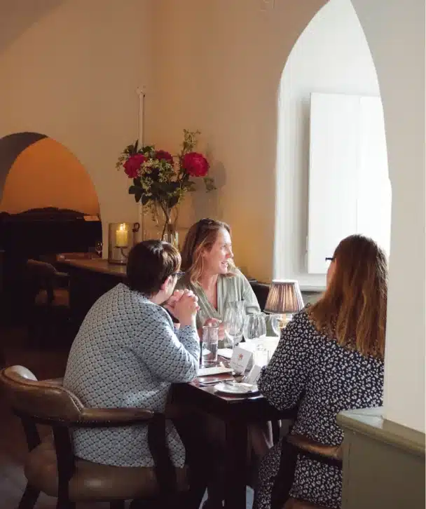 Three women sit at a table in Nugents Hall, Malahide Castle, talking and smiling. The cozy restaurant setting includes glasses, plates, and a vase with flowers near a softly lit window in the background.