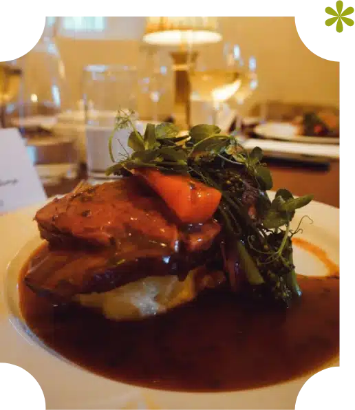 A plated gourmet meal at Nugents Hall Malahide Castle features slices of beef with brown sauce, mashed potatoes, broccolini, and carrots, garnished with microgreens. Wine glasses and a lamp are blurred in the background.