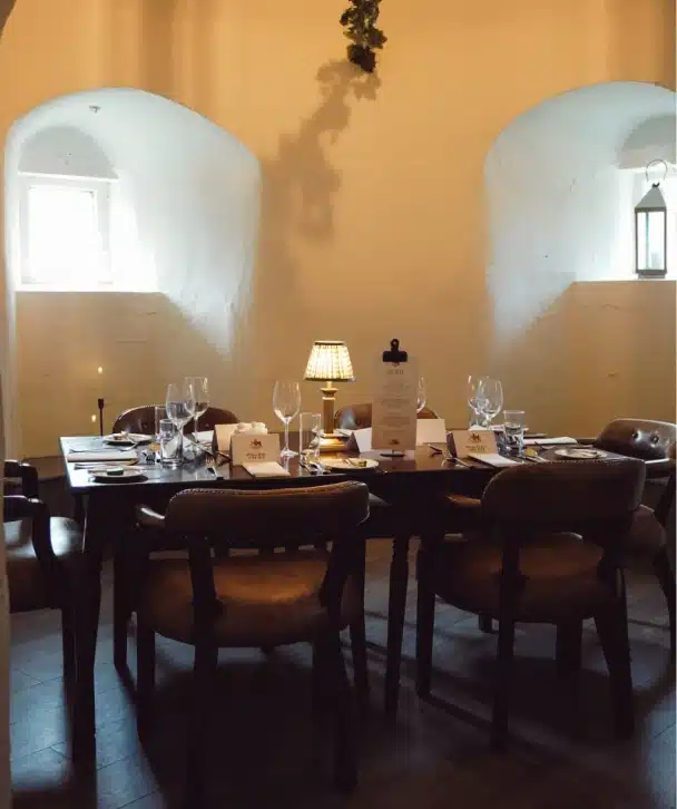 A cozy, elegantly set dining table with six chairs graces Nugents Hall, Malahide Castle. Wine glasses, menus, and a small lamp adorn the table as light streams through arched windows, creating a warm, intimate atmosphere.