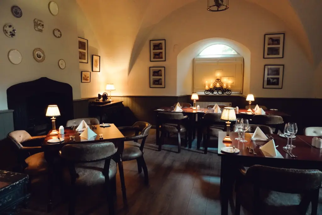 A cozy, dimly lit restaurant with wooden floors, set tables, cushioned chairs, and framed pictures evokes the inviting charm of a Hunt Dinner Party at Malahide Castle, with lamps casting warm light and a window at the back.