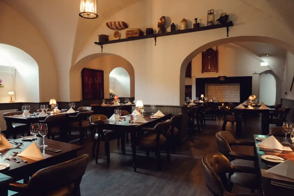 A cozy, upscale restaurant with arched ceilings, cushioned chairs, and warm lighting evokes the elegance of a Hunt Dinner Party at Malahide Castle; decorative items line a high shelf above the neatly set wooden tables in the empty dining area.