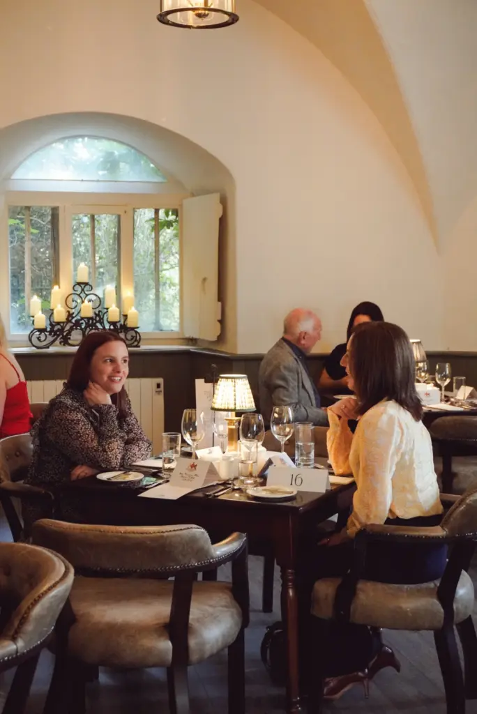 People sit at elegant tables in a softly lit restaurant with arched ceilings and candles on the windowsill, evoking the charm of a Hunt Dinner Party at Malahide Castle. Two women face each other in lively conversation at a table in the foreground.