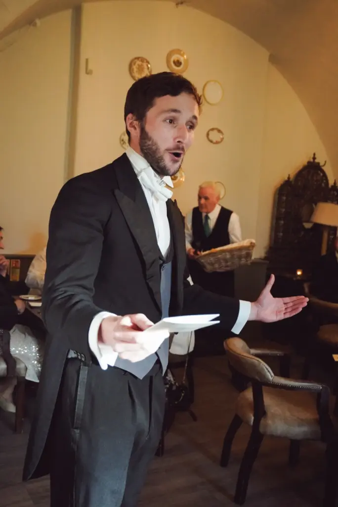 A man in formal attire sings passionately with open arms, holding a piece of paper at the Hunt Dinner Party at Malahide Castle. He stands in an elegant room with seated guests, ornate wall decor, and a server in the background.