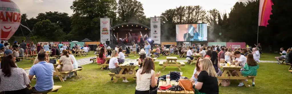 People sit at picnic tables on a grassy field, watching a live outdoor concert on stage. A large screen displays the performers, and trees surround the area, creating a relaxed festival vibe typical of Summer Concerts at Malahide Castle.