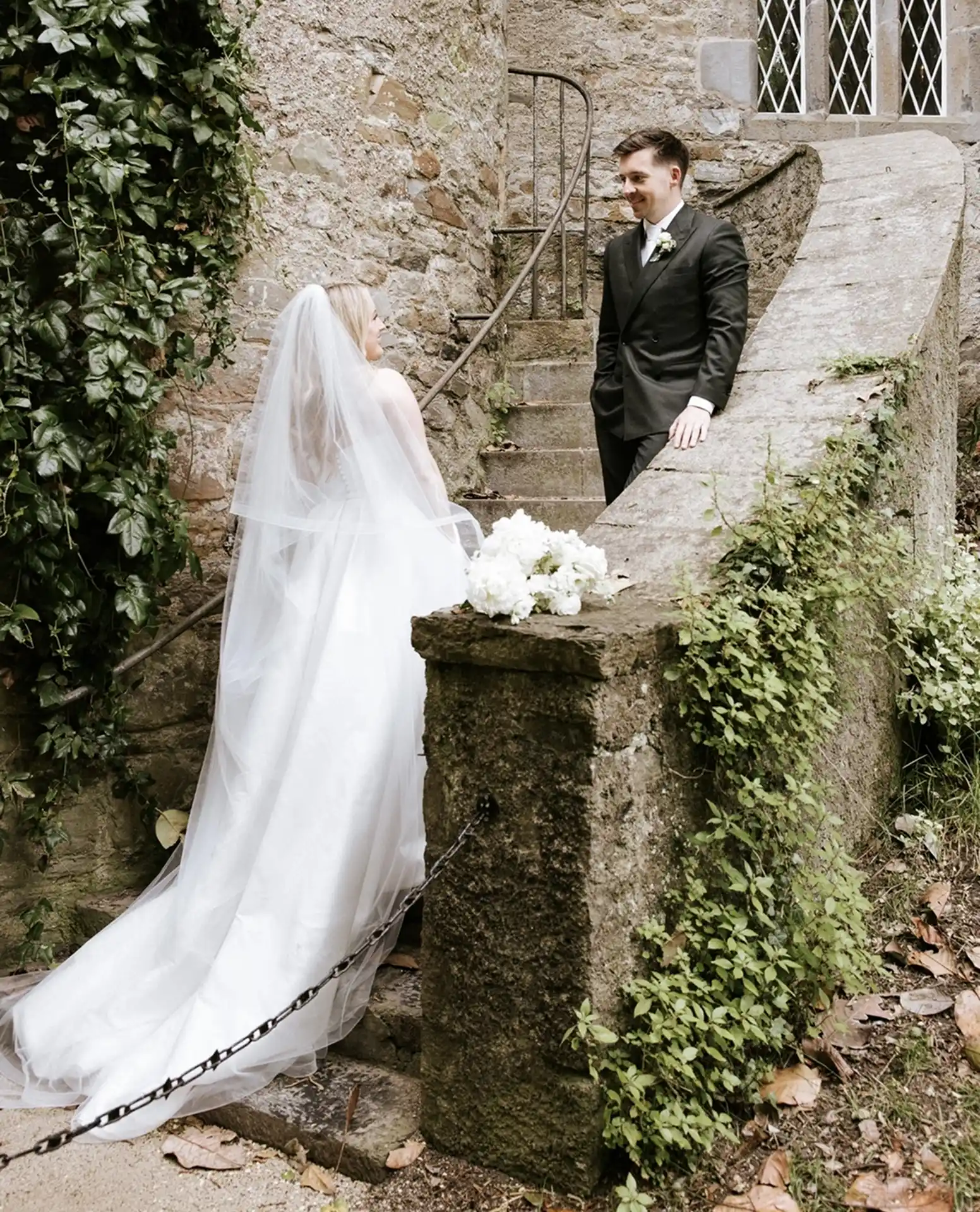 A bride in a white gown and veil stands at the bottom of stone steps at Malahide Castle, looking up at a groom in a black suit who is smiling down at her—an enchanting spot perfect for private hire.