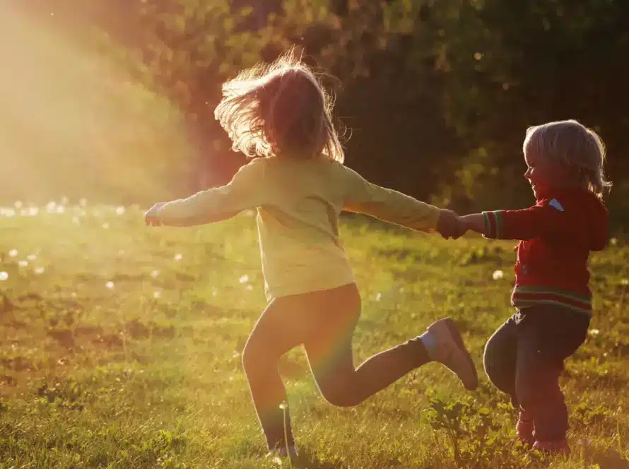 Two young children holding hands run through a sunlit grassy field, with trees in the background and warm sunlight streaming in from the left.