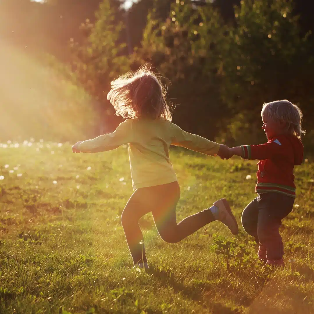Two young children holding hands run through a sunlit grassy field, with trees in the background and warm sunlight streaming in from the left.