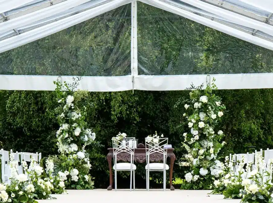 A wedding ceremony setup under a clear tent with rows of white chairs, floral arrangements along the aisle, and two chairs at the altar surrounded by greenery and white flowers—an ideal scene for Private Hire Malahide Castle.