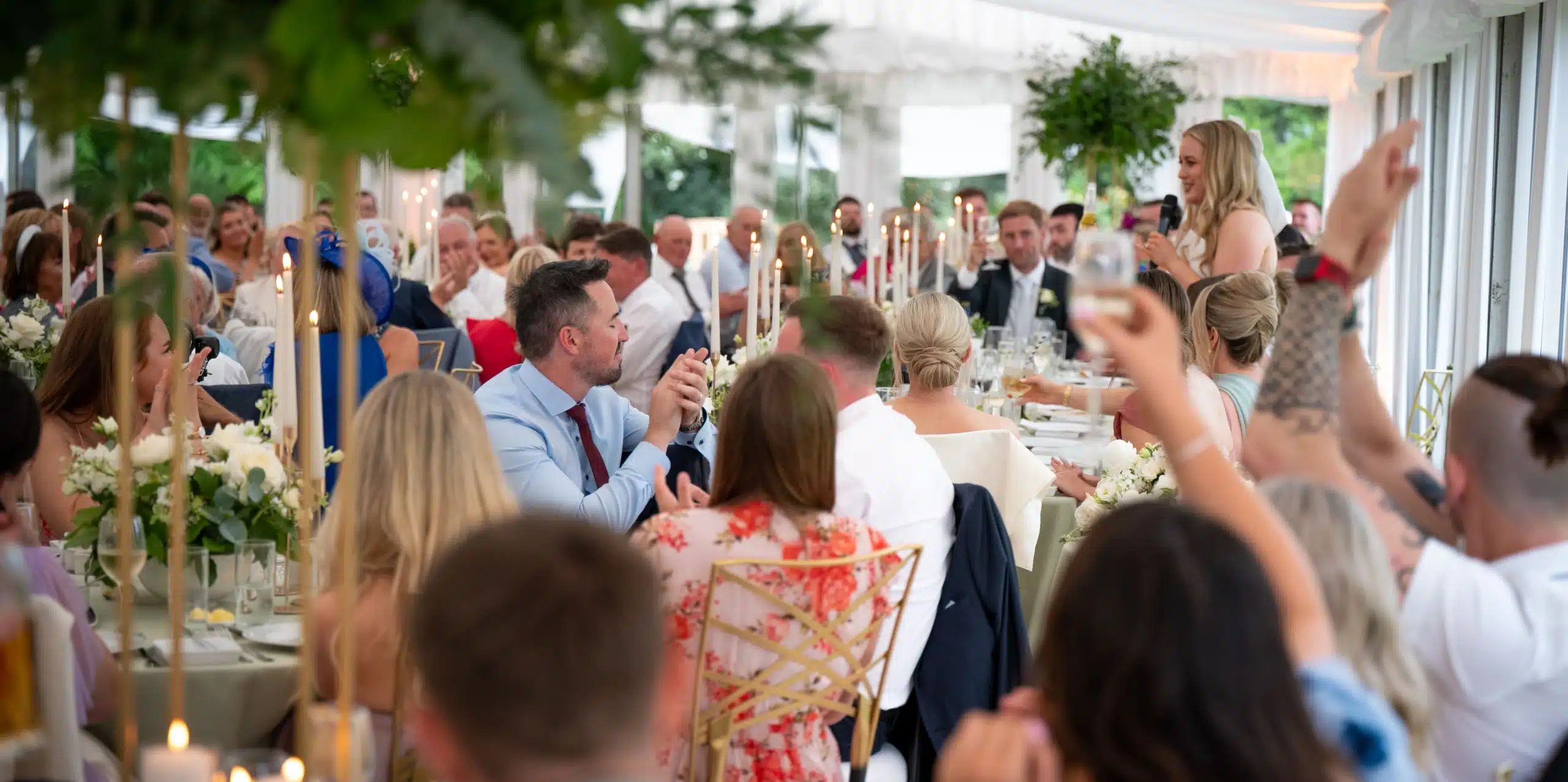Guests seated at elegantly decorated tables in a bright, white Malahide Castle marquee hire raise glasses in a toast while a woman stands and gives a speech at the front during a wedding reception.