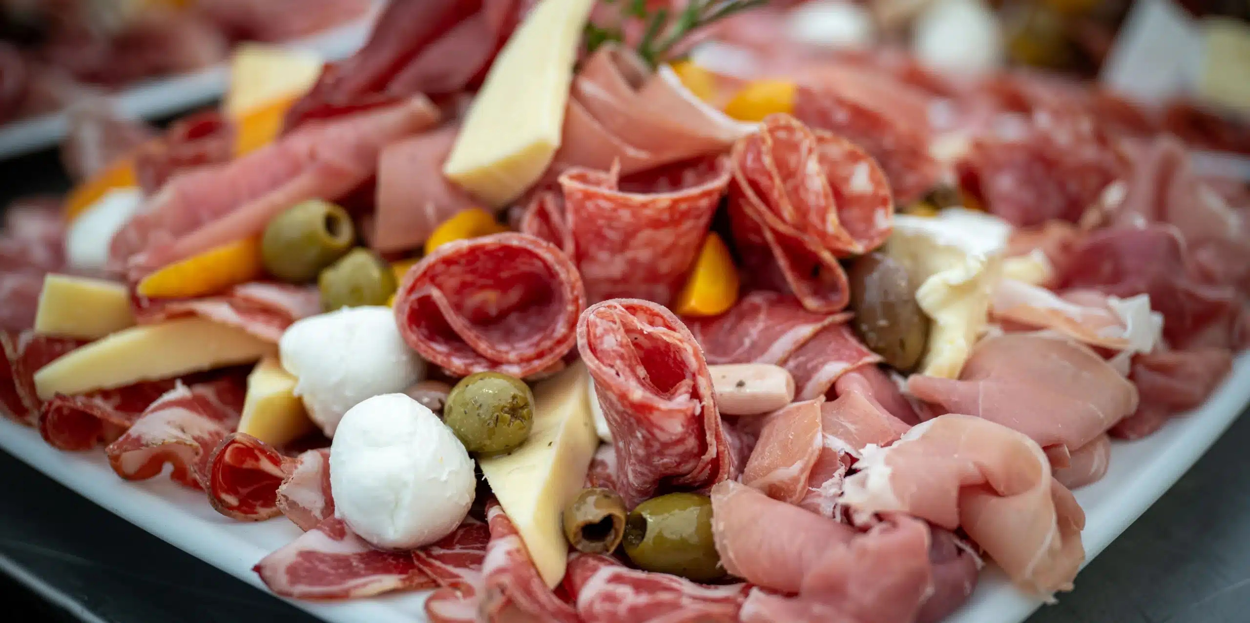 A close-up of a charcuterie platter with slices of cured meats, salami, prosciutto, cheese, mozzarella balls, green olives, and yellow peppers, perfect for enjoying at a Malahide Castle marquee hire event.