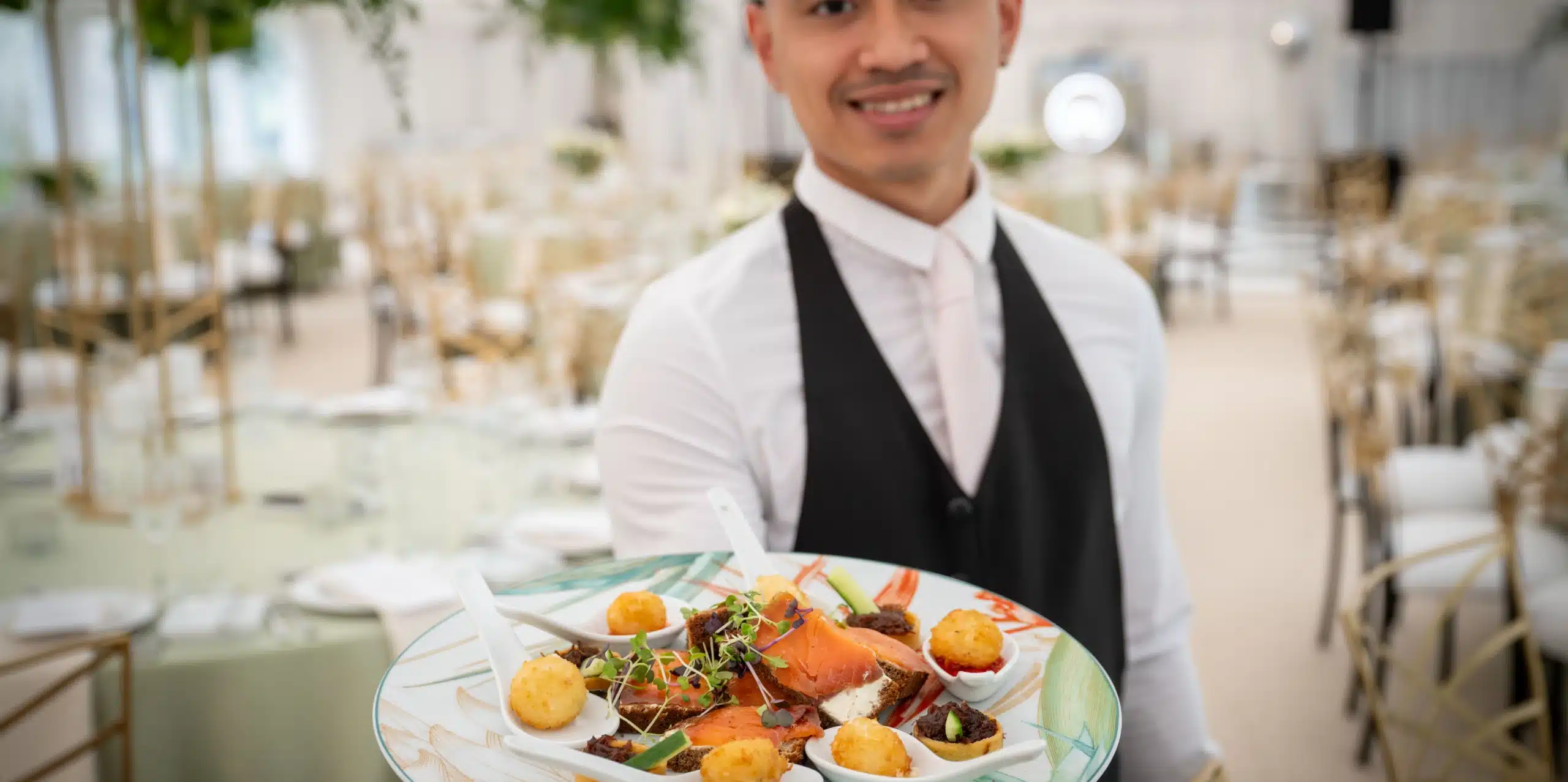 A smiling waiter in formal attire holds a platter of assorted appetizers, including salmon, meat, and fried bites, in an elegant dining setting with round tables—perfect for events with Malahide Castle marquee hire.
