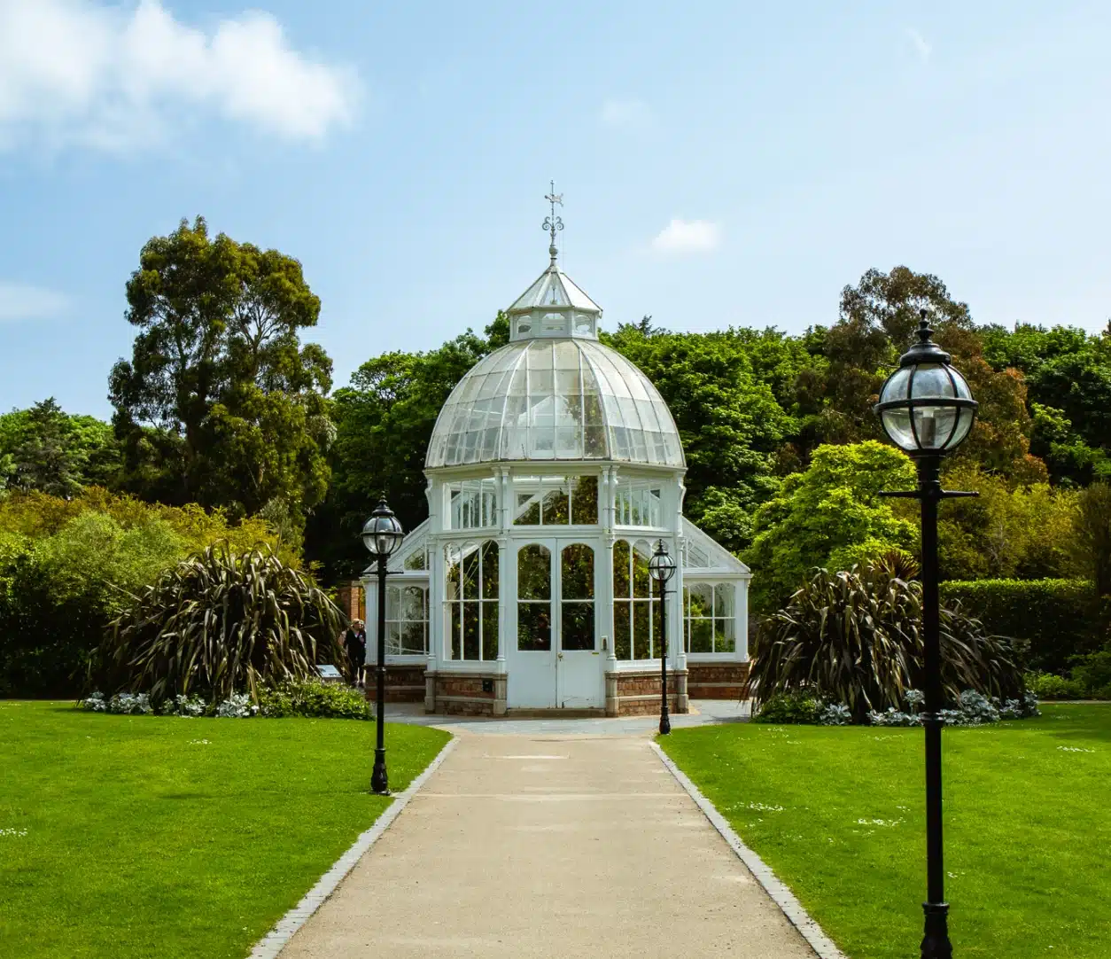 A white, glass-domed greenhouse sits at the end of a paved path near Malahide Castle marquee hire, surrounded by lush green lawns, trees, and two black lamp posts under a bright blue sky.