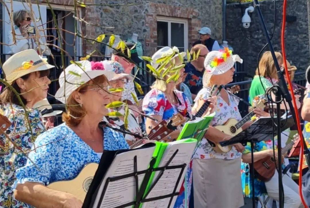 A group of women in colorful floral outfits and hats play ukuleles and sing outdoors, bringing to life melodies from A Traitorous Tale. They stand before a stone building, with music stands and a microphone in view.