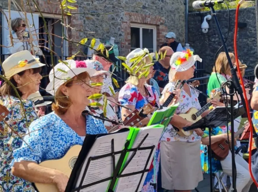 A group of women in colorful floral outfits and hats play ukuleles and sing outdoors, bringing to life melodies from A Traitorous Tale. They stand before a stone building, with music stands and a microphone in view.