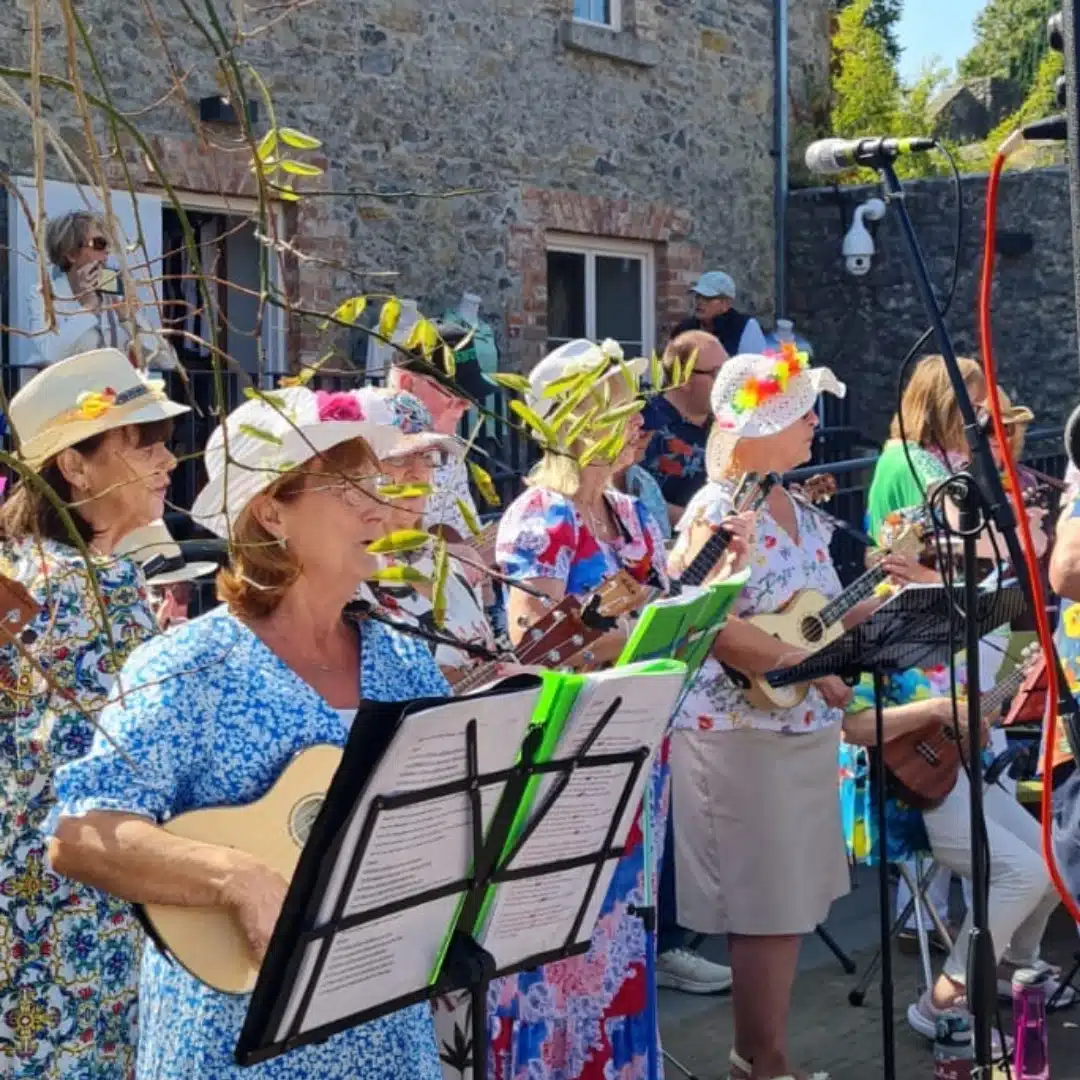 A group of women in colorful floral outfits and hats play ukuleles and sing outdoors, bringing to life melodies from A Traitorous Tale. They stand before a stone building, with music stands and a microphone in view.