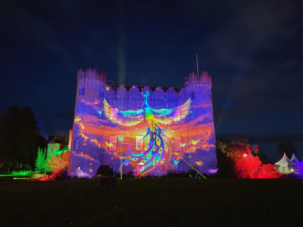 A castle wall is illuminated at night with a vibrant projection of a glowing phoenix and shimmering purple-orange clouds, capturing the magic of Wonderlights - Winter's Brightest Tradition.