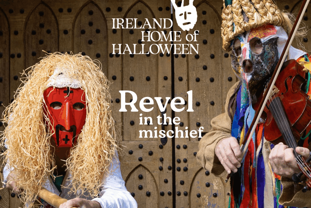 Two people wearing colorful, traditional Irish Halloween costumes and masks stand in front of a wooden door. One plays a fiddle, the other holds a stick.