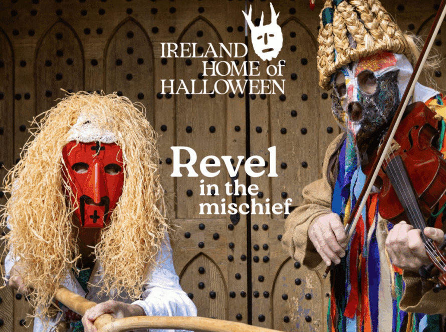 Two people wearing colorful, traditional Irish Halloween costumes and masks stand in front of a wooden door. One plays a fiddle, the other holds a stick.