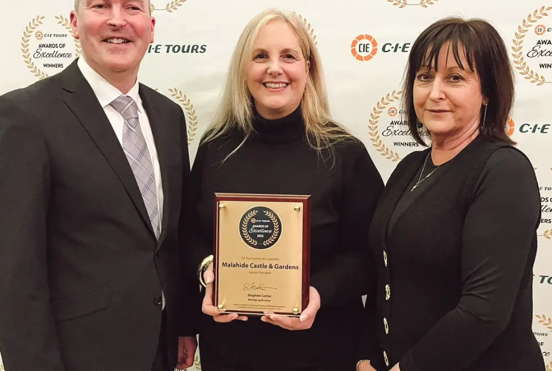 Three people in formal attire pose for a photo. The woman in the center holds a plaque that reads "Malahide Castle & Gardens." The "CIE Tours" awards backdrop hints at achievements.