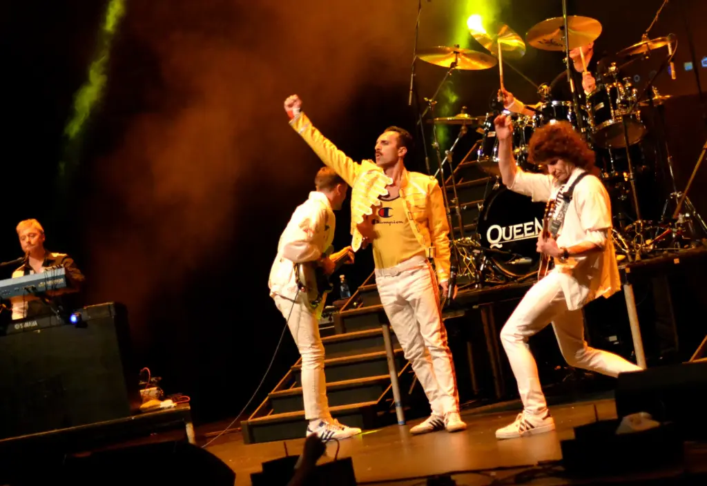 A rock band performs energetically on stage; the lead singer in a yellow jacket raises his fist, flanked by two guitarists and a keyboard player, whilst bright lights illuminate “Queen by Moonlight” displayed on the drum kit.
