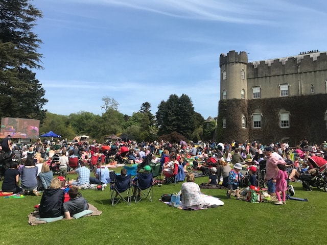 A large crowd sits on blankets and chairs on a lawn, enjoying Summer Sessions at the Castle near a historic stone castle under a blue sky. Trees and greenery surround the lively outdoor event.