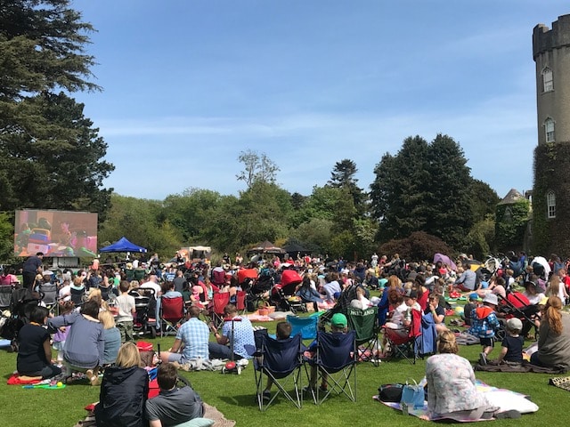 A large crowd relaxes on blankets and deckchairs at Summer Sessions at the Castle, enjoying a sunny day whilst watching a large screen set up on the grass, surrounded by trees and the iconic castle building.