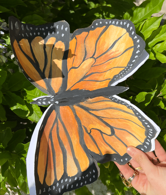 A hand holds a large, realistic paper butterfly painted orange, black, and white—its delicate wings glowing like a Queen by Moonlight—against a background of sunlit green leaves.