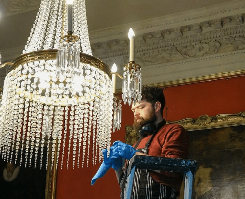 During National Heritage Week, a person standing on a ladder wears an apron and blue gloves, carefully cleaning a large, ornate crystal chandelier in a room with red walls and elaborate ceiling mouldings.