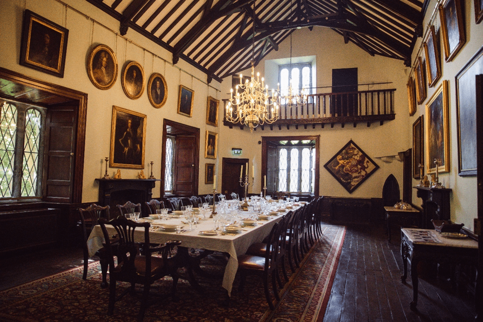 A long dining table set for a formal meal is in an ornate hall with high vaulted ceilings, chandeliers, paintings, and large windows—perfect for celebrating National Heritage Week with elegant place settings and dark wood chairs neatly arranged.