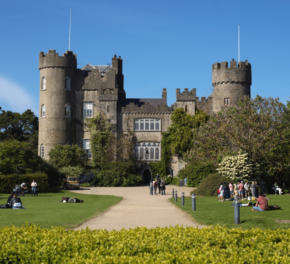 A large stone castle with round towers and battlements stands under a clear blue sky, surrounded by gardens. During National Heritage Week, several groups of people are gathered on the lawn in front of the castle.
