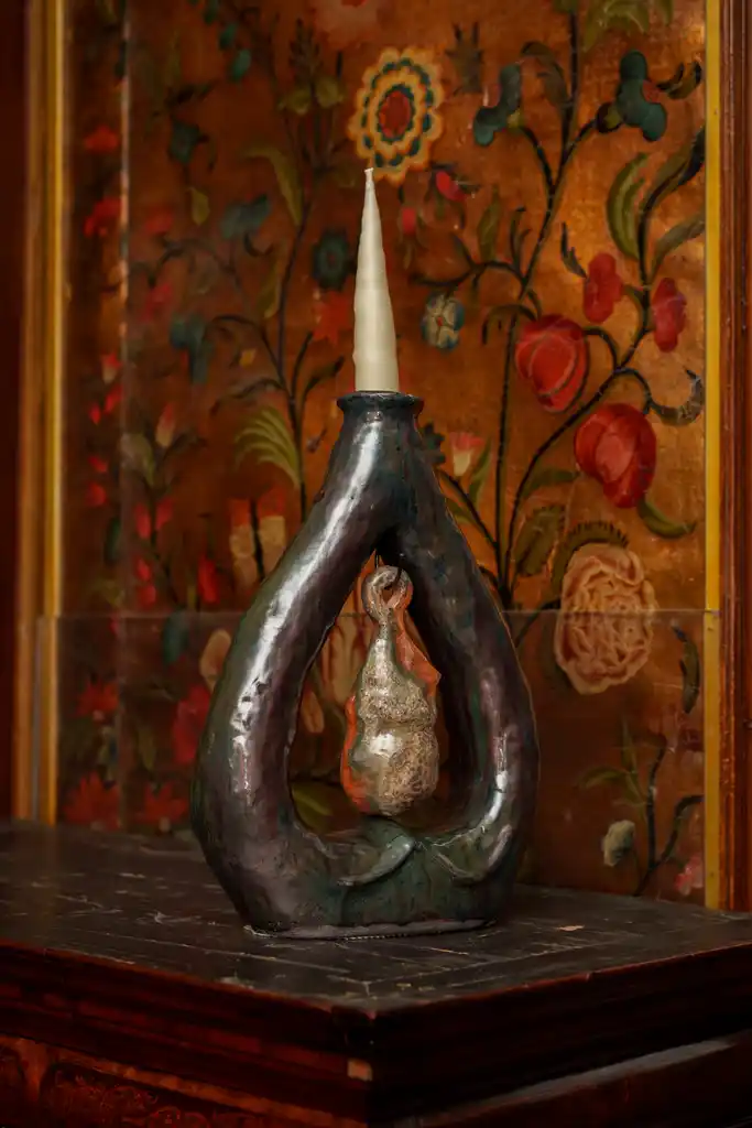 A sculptural candleholder with a white candle, featuring a looped, organic metal form holding a small hanging object, stands on a dark surface against a floral-decorated golden background—an elegant piece perfect for illuminating the ambience of Culture Night 2025.