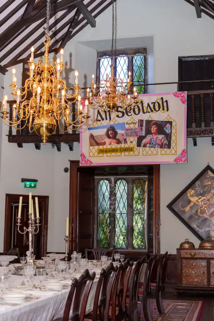 A formal dining room with chandeliers, candelabras, and a long set table. A large colourful banner with photos and text, including “An Seisiún” and a reference to Culture Night 2025, hangs above dark wooden doors.