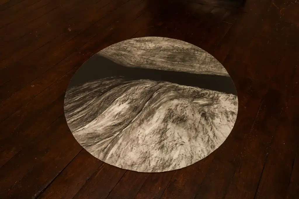 A round artwork on a wooden floor depicts a mountainous landscape in greyscale, divided by a dark, horizontal shape that resembles a river or chasm cutting through the scene. This striking piece will be featured as part of Culture Night 2025, inviting viewers to contemplate the dramatic contrasts and stories within the landscape.