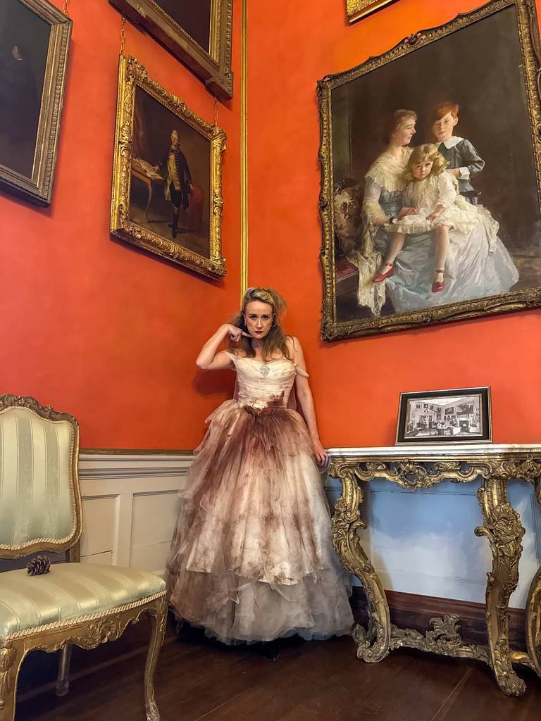 A woman in a vintage gown stands by an ornate table in a lavish room with orange walls, gold-framed paintings, and an upholstered chair, as if awaiting the next chapter of A Treacherous Tale to unfold.