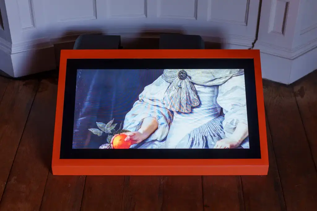 A digital screen in an orange frame displays a classical painting of a person holding an orange fruit, resting on a wooden floor against a white panelled wall—an inviting scene that sets the tone for Culture Night 2025.