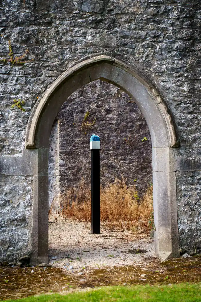 A stone archway in a grey brick wall frames a small black post with a blue and white object on top, surrounded by dry grass and another stone wall in the background—an intriguing scene that could easily be part of the ambience for Culture Night 2025.