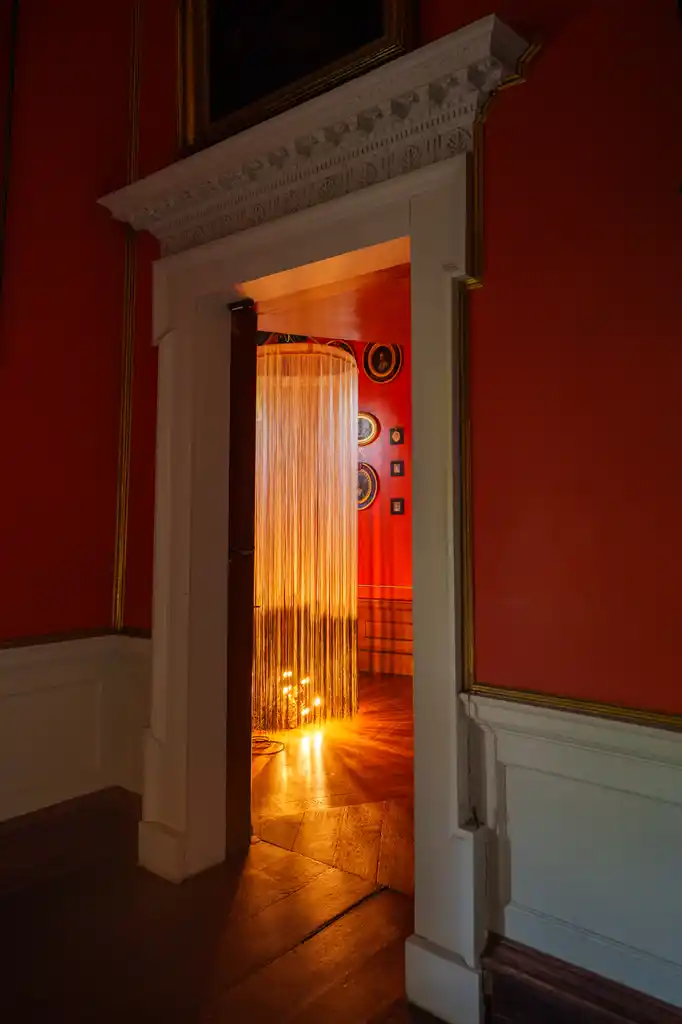A warmly lit room with red walls and framed art is seen through an open door, inviting guests to experience Culture Night 2025. A curtain of hanging strings glows with light at its base, casting reflections on the wooden floor.