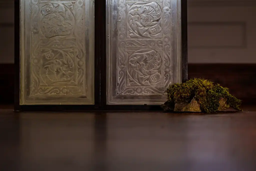 A piece of moss-covered rock sits on a wooden floor next to ornate, engraved metal panels with bird and floral designs—evoking the natural beauty and artistic heritage celebrated during Culture Night 2025.