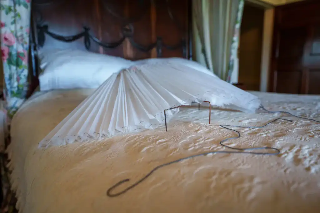 A white wire and paper bonnet sits on a patterned bedspread in a vintage bedroom, with a wooden headboard and pillows in the background, evoking the timeless charm of traditions celebrated during Culture Night 2025.