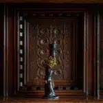 A small, dark bronze sculpture with yellow accents stands in front of an ornately carved wooden panel featuring intricate patterns and rich brown tones, quietly evoking the mysterious ambience of A Traitorous Tale.