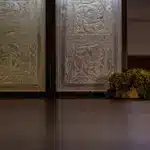 A small patch of moss sits on the floor next to a decorative metal panel with intricate floral patterns, against a dimly lit wall—a quiet scene that could belong in A Treacherous Tale.