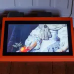 A digital screen in an orange frame, placed on a wooden floor, displays part of a classical painting showing a person in white clothing holding a red apple—a scene reminiscent of “A Treacherous Tale.”.