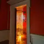 A doorway opens to a room with a floor-to-ceiling lighted string curtain, evoking an air of mystery reminiscent of A Treacherous Tale. Warm light glows behind the curtain, and several circular decorations hang on the far wall. The walls are painted red with white trim.