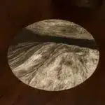 A round artwork resembling a black and white landscape with a river sits on a dark wooden floor, evoking the mood of A Traitorous Tale. The drawing features textured hills and a winding waterway through the centre, suggesting secrets hidden within its shadowy contours.