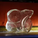 Translucent, irregularly shaped sculpture with red and white swirls sitting on a reflective surface, against an orange and dark blue background—an evocative scene reminiscent of A Treacherous Tale.
