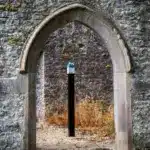 A stone archway in an old wall frames a modern black pole with a blue-topped fitting, standing on gravel with dry weeds and another stone wall in the background—an evocative scene reminiscent of A Traitorous Tale unfolding between past and present.
