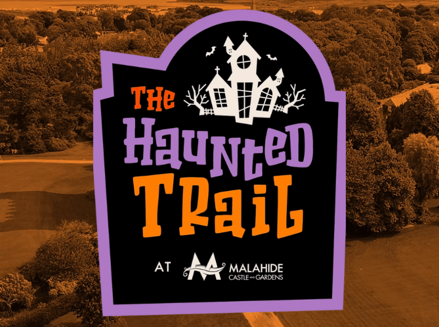 A Halloween-themed sign reading “The Haunted Trail at Malahide Castle & Gardens” stands prominently against an orange-tinted aerial view of a park with trees and open grassy areas, evoking the mysterious allure of The Haunted Trail.