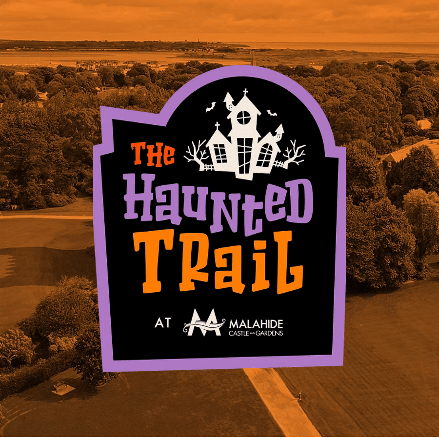 A Halloween-themed sign reading “The Haunted Trail at Malahide Castle & Gardens” stands prominently against an orange-tinted aerial view of a park with trees and open grassy areas, evoking the mysterious allure of The Haunted Trail.